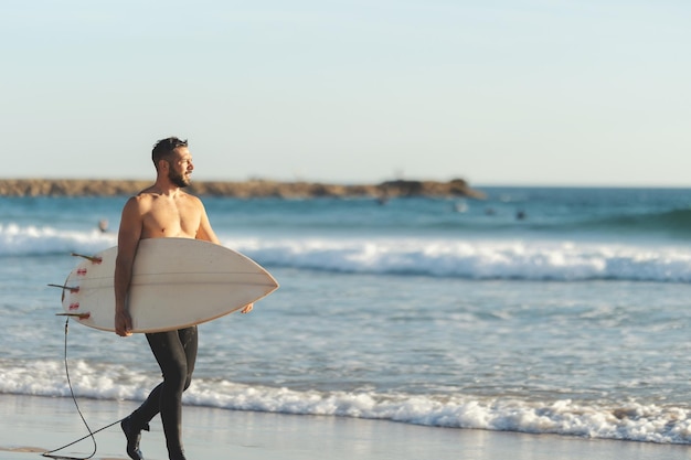 A smiling man surfer with naked torso walking on the seashore