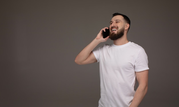 Uomo sorridente in maglietta che parla dallo smartphone e che esamina la macchina fotografica sopra fondo grigio.