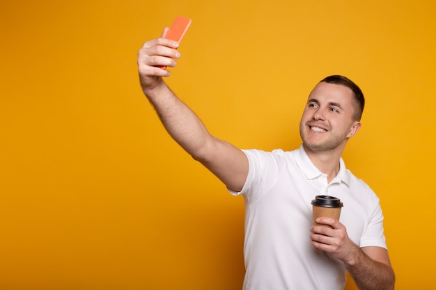 Smiling man taking selfie on smartphone with coffee to go on yellow