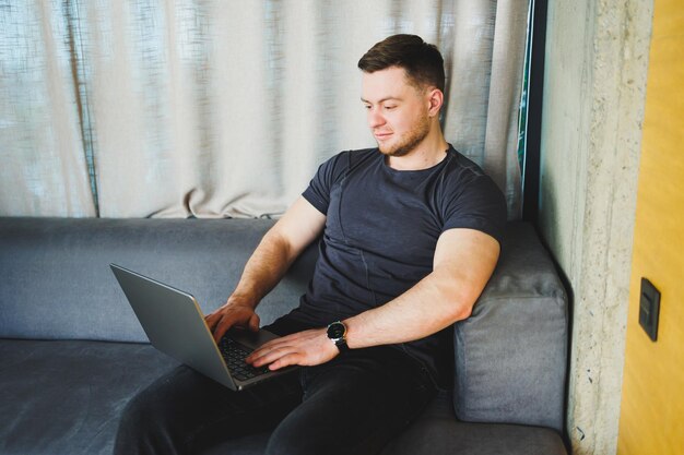 Smiling man in tshirt sitting on sofa typing on netbook working remotely on startup as freelancer looking at laptop and smiling