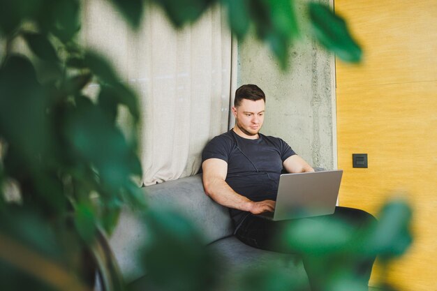 ソファに座ってネットブックに入力するTシャツを着た笑顔の男性が、フリーランサーとして起動時にリモートで作業し、ノートパソコンを見て微笑んでいる