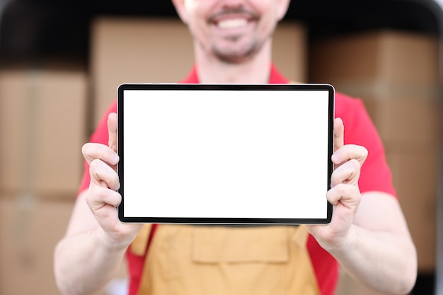 Улыбающийся человек в форме держит планшет с белым экраном на фоне картонных коробок