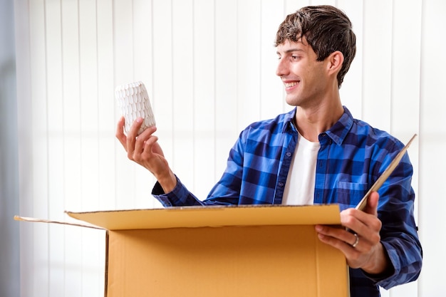 Uomo sorridente che disimballa il pacco atteso con in mano un vaso seduto con una tenda sullo sfondo cliente felice soddisfatto che apre la scatola di cartone con l'ordine del negozio online buon servizio di consegna spedizione