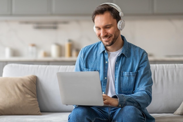 Smiling Man Uses Laptop While Wearing Headphones on a Couch