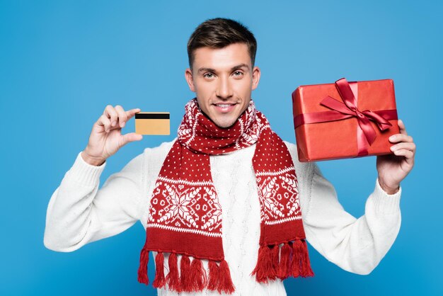 Smiling man wearing scarf and sweater showing wrapped present and credit card isolated on blue