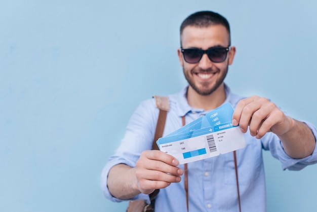 Occhiali da sole da portare sorridenti dell'uomo che mostrano biglietto aereo su priorità bassa blu