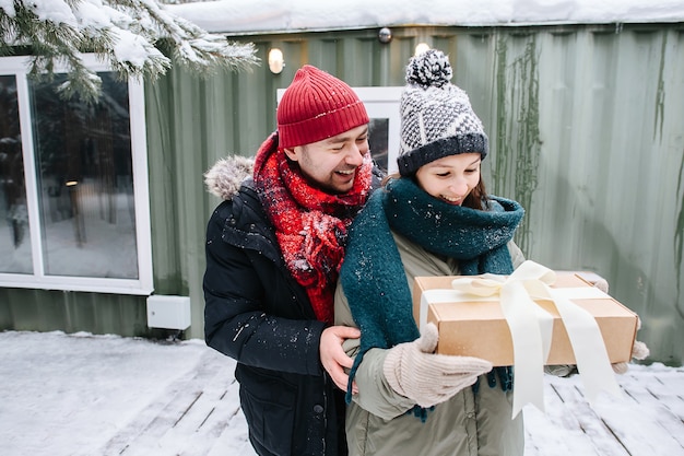 Smiling man in winter clothes gifted woman with a present in a\
bow-tied box. standing in front of a small snowy house.