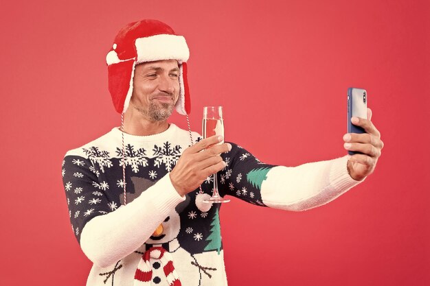 Foto uomo sorridente in maglione invernale e cappello che fa selfie su smartphone con champagne xmas