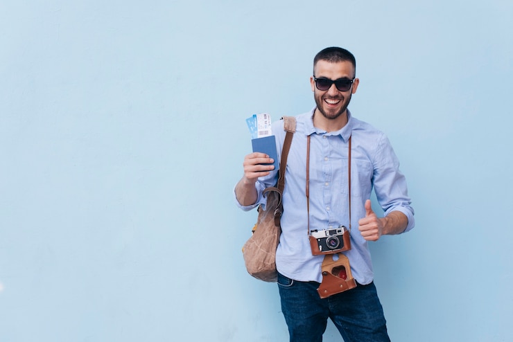 Photo smiling man with camera around his neck holding air ticket and showing thumb up gesture standing near blue wall