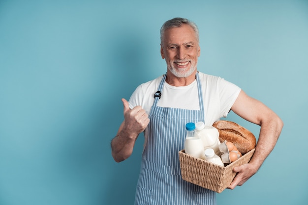 Smiling man with gray hair and a beard holds a basket of food and points his thumb at the copy space