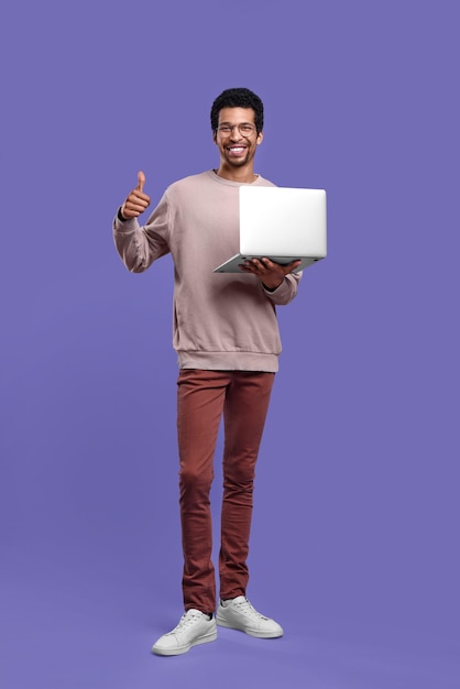 Smiling man with laptop showing thumb up on purple background