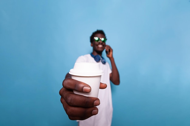 Smiling man with smartphone showing cup of coffee to camera. Casual adult wearing sunglasses and headphones holding drink while talking on phone call and feeling carefree. Person having fun