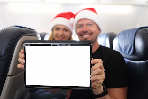 Smiling man and woman in santa claus hats hold tablet with blank white screen in aircraft cabin