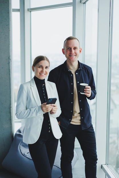 Smiling man and woman standing near window with gadgets in modern transparent office Conversation of business colleagues in an informal setting