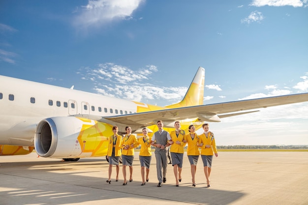 Smiling man and women airline workers wearing aviation uniform while walking near passenger plane at airport