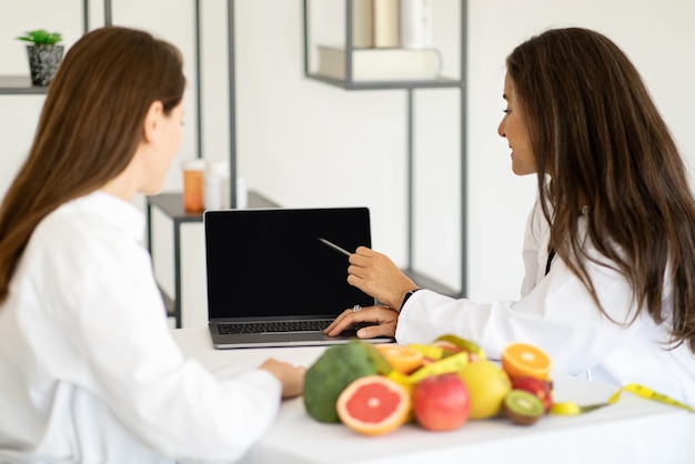 Smiling mature caucasian doctor nutritionist consulting young woman shows diet plan at laptop with