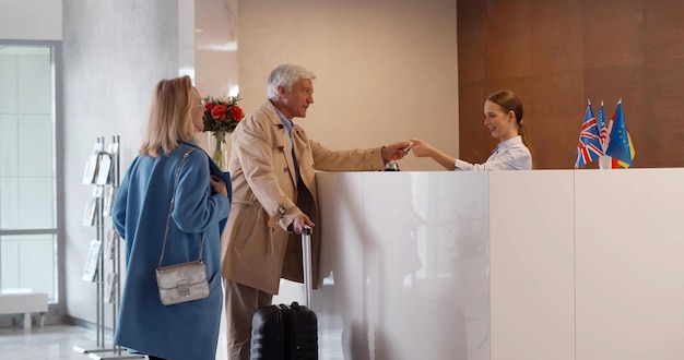 Foto coppia matura sorridente alla cassa e parla con l'addetto alla reception nella hall dell'hotel