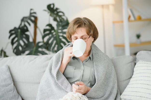 Smiling mature elder 65s woman sitting relaxing with cup of tea coffee Senior mid age stylish look woman with eyeglasses portrait with cup looking away at modern home