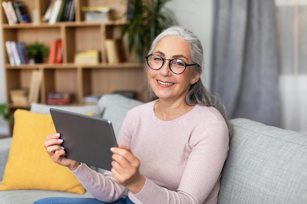 眼鏡をかけた笑顔の成熟したヨーロッパの白髪の女性が、リビングルームのインテリアでタブレットチャットビデオを見る