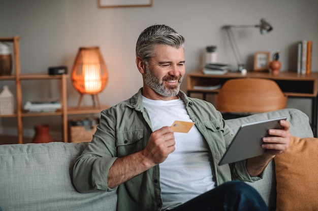 Smiling mature european man with beard with credit card looks at tablet and pay in living room