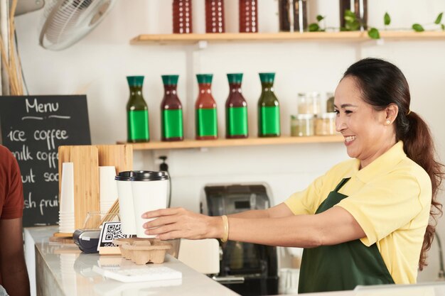 Smiling mature female coffeeshop owner giving customer two big cup of take out coffee