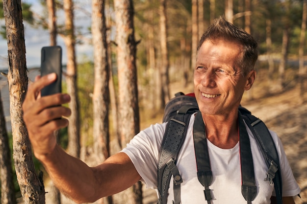 Smiling mature male is walking in nature with backpack and using smartphone for taking photo