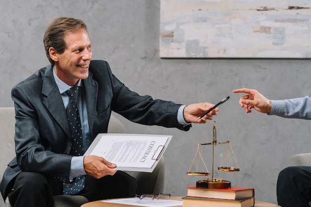 Улыбаясь зрелый мужчина-юрист, держащий контракт, вручая ручку своему клиенту