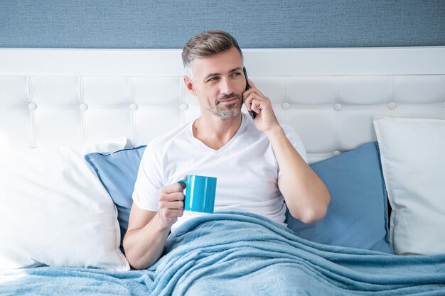 Smiling mature man drinking coffee in bed and talking on phone