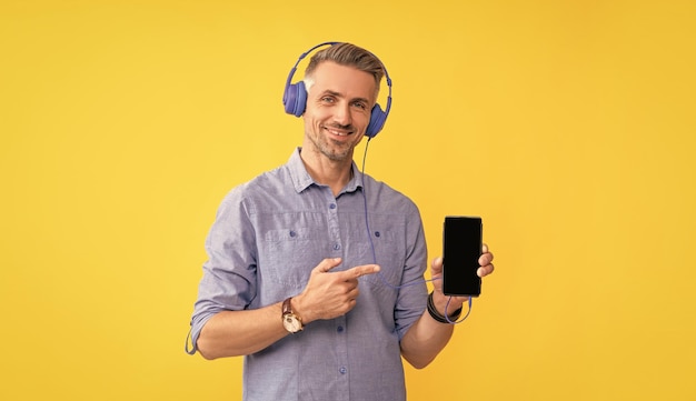 Smiling mature man listen music in headphones pointing finger on phone presentation