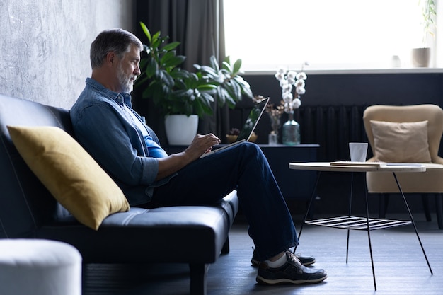 Smiling mature man sitting on a sofa typing on a laptop in a living room. Working from home in quarantine lockdown. Social distancing Self Isolation