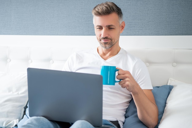 Smiling mature man using computer in bed with coffee