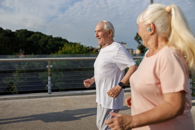Улыбающиеся зрелые мужчина и женщина с хвостиком вместе бегают по пешеходному мосту в летний день