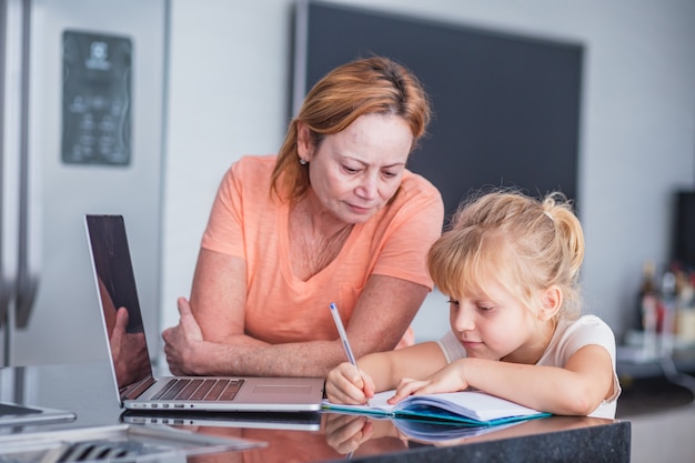 Smiling mature mother helps her daughter in preparing chores at home. Online education concept.