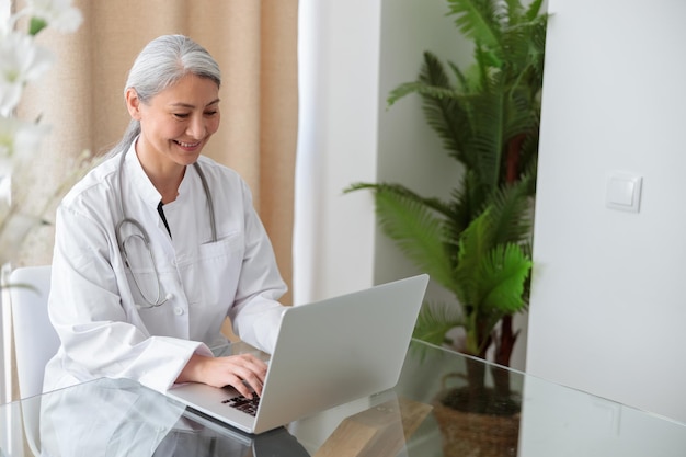 Smiling mature physician lady in white coat working on laptop