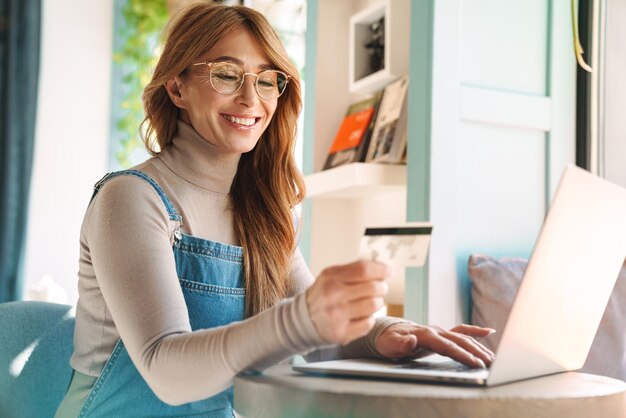 Sorridente donna matura in occhiali tenendo la carta di credito durante l'utilizzo su laptop in una stanza luminosa