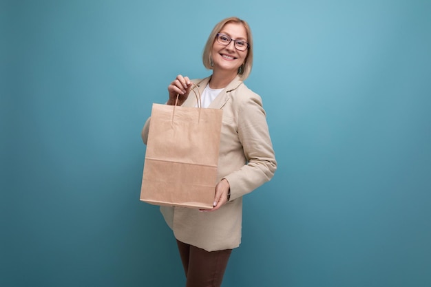 Smiling mature woman in jacket holding craft shopping bag on studio background with copy space