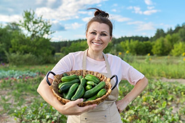 Улыбающаяся зрелая женщина с корзиной свежих сорванных огурцов на заднем дворе, выращивающая натуральные органические овощи, хобби и досуг, концепция здорового питания