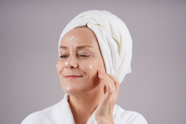 Smiling mature woman with white towel on her head applying cream to her face. Facial care concept. Isolate on gray wall