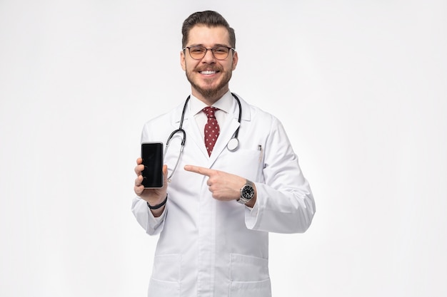 Smiling medical doctor holding smartphone. Isolated over white wall