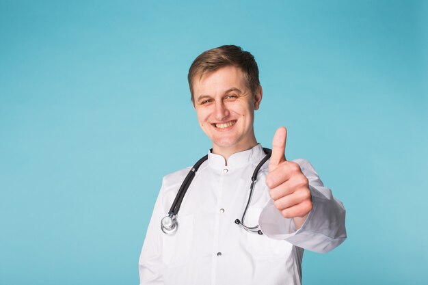 Uomo sorridente del medico con lo stetoscopio che mostra i pollici su sopra fondo blu con lo spazio della copia