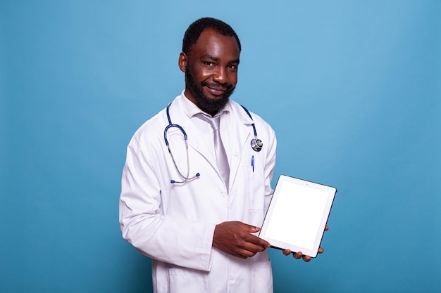 의료 개념 사진을 위한 흰색 화면 디지털 태블릿을 제시하는 청진기를 들고 웃고 있는 의사. 모형 디스플레이가 있는 디지털 터치스크린 컴퓨터를 들고 실험실 가운을 입은 친절한 의료진.