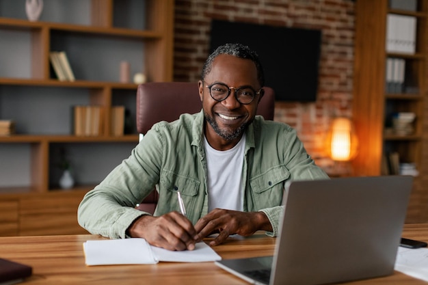 Улыбающийся афроамериканец средних лет в очках работает на ноутбуке за столом, делает заметки за столом в офисе дома