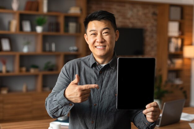 Smiling middle aged chinese man ceo manager show finger at\
tablet with empty screen in home office interior