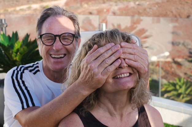 Smiling middle aged couple family in outdoor terrace have fun man covering the female eyes with hand