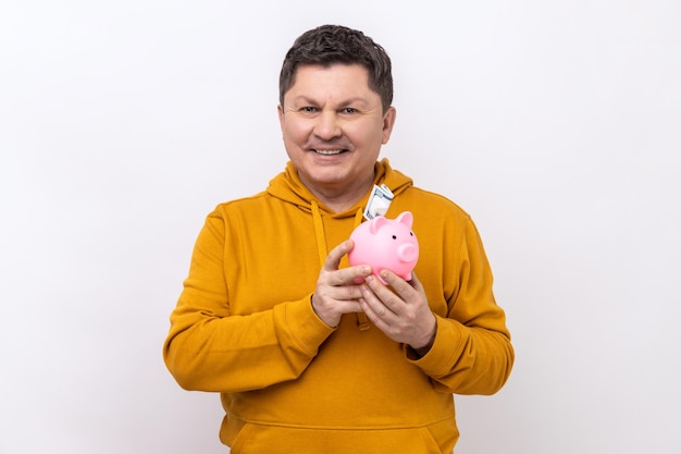 Smiling middle aged man holding piggy bank with banknote in hands being happy of savings