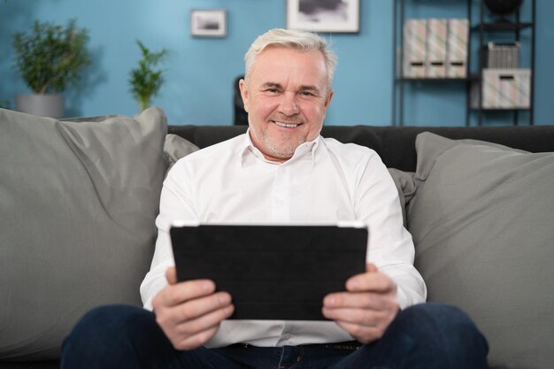 Smiling middle aged old man relaxing holding digital tablet reading e book sit on sofa at home