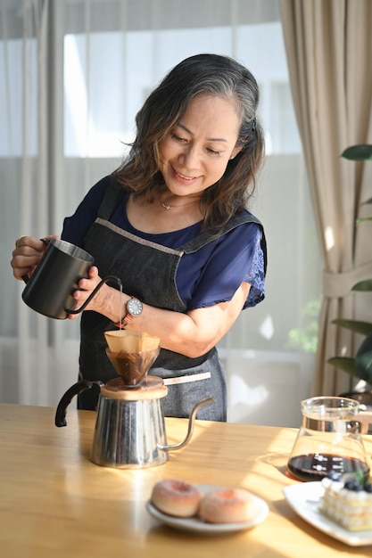 Smiling middle aged standing at kitchen table brewing drip coffee and enjoys her morning