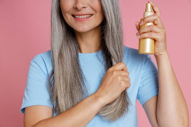 Foto sorridente donna di mezza età applica il prodotto spray sulla ciocca di capelli grigi in piedi su sfondo rosa in studio closeup. stile di vita di bellezza matura