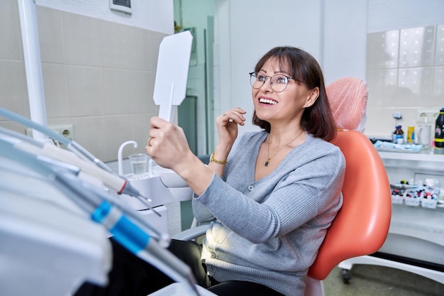 Smiling middle aged woman in dental chair with mirror looking at her teeth