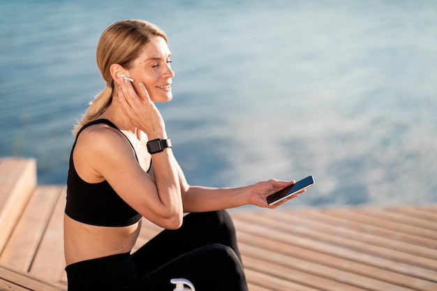 Smiling Middle Aged Woman In Earphones Resting With Smartphone After Training Outdoors